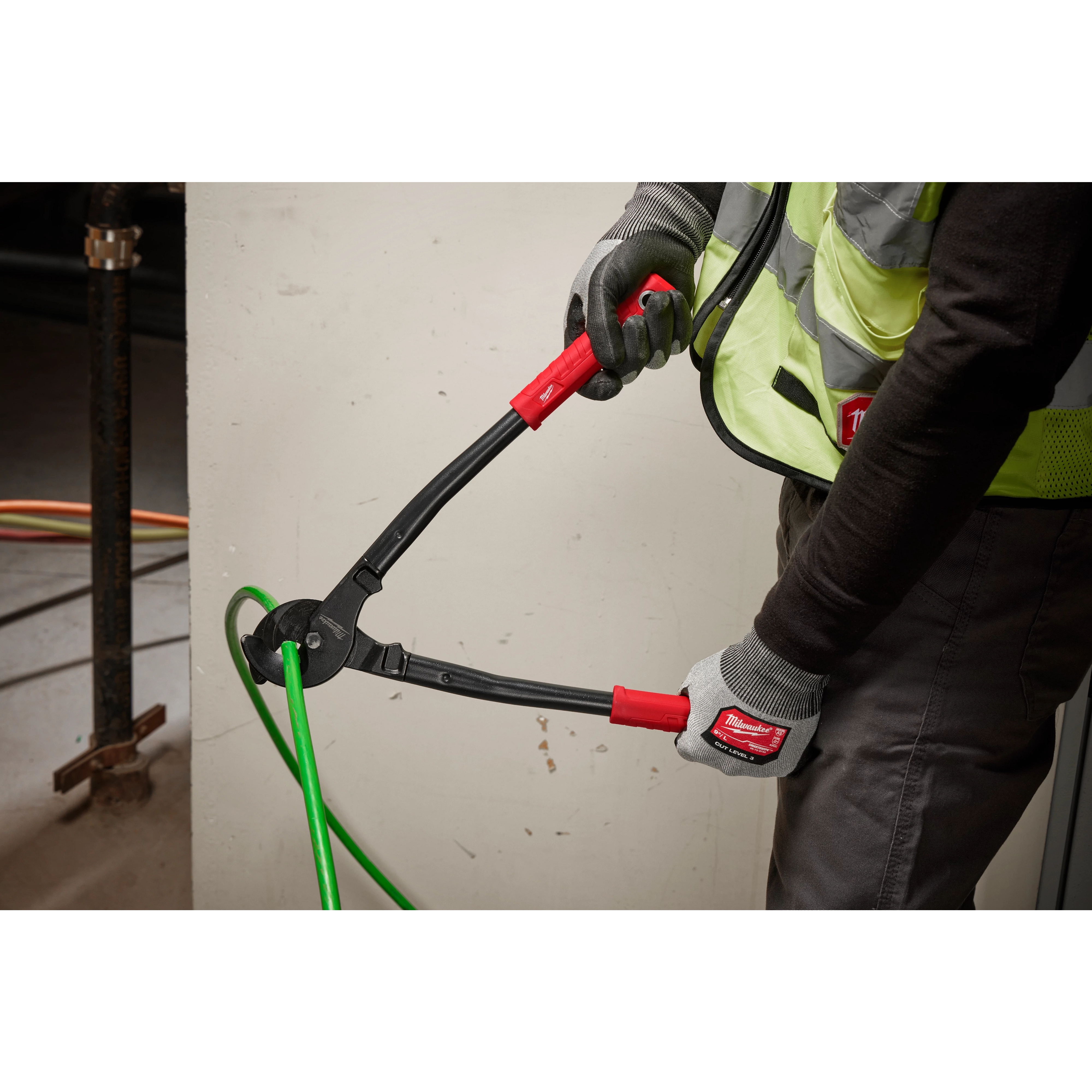 A worker uses 17" Utility Cable Cutter with red handles to cut a thick green cable, wearing gloves and a high-visibility vest.