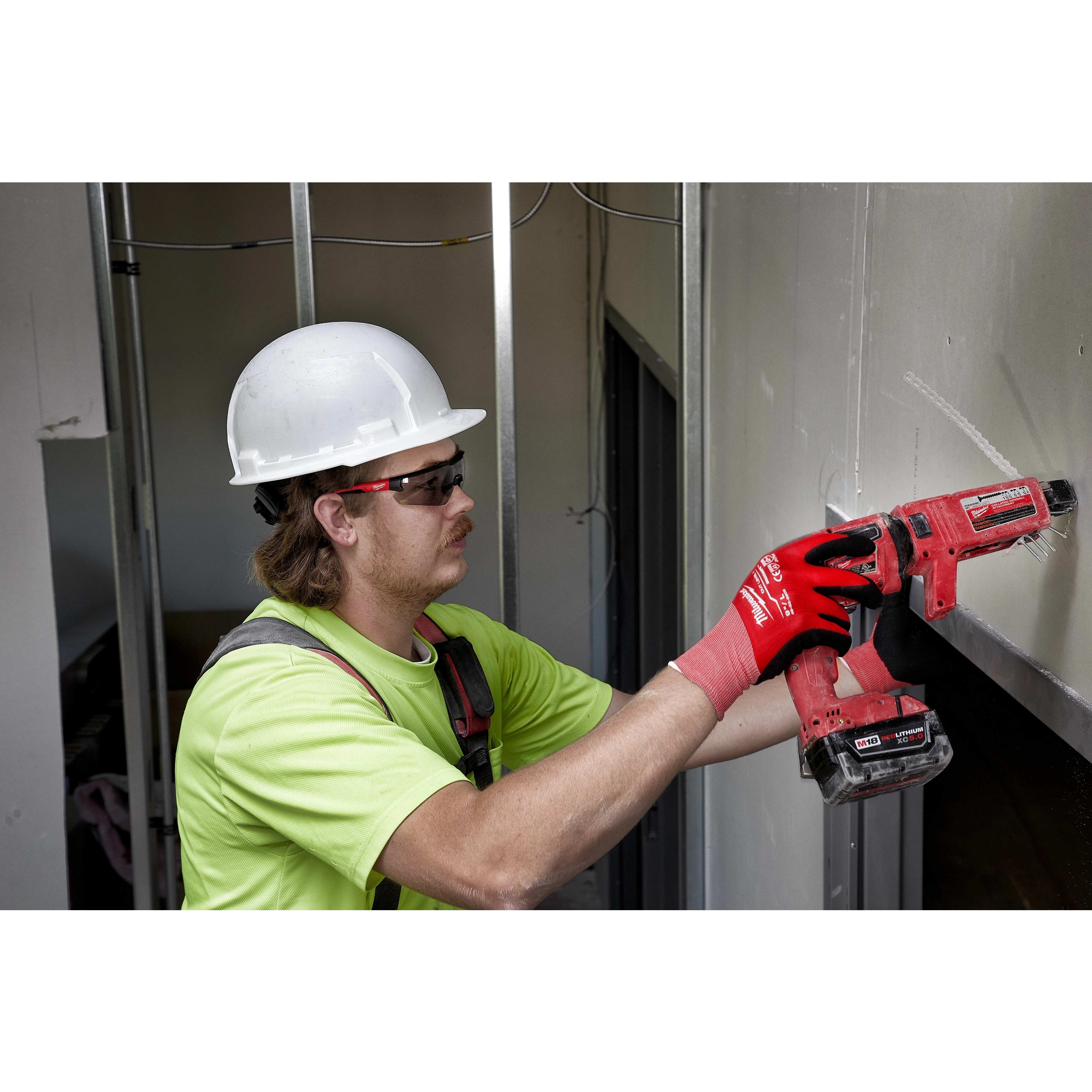 A worker wearing a hard hat, gloves, and Safety Glasses - Gray Anti-Scratch Lenses uses a red power drill to install drywall.