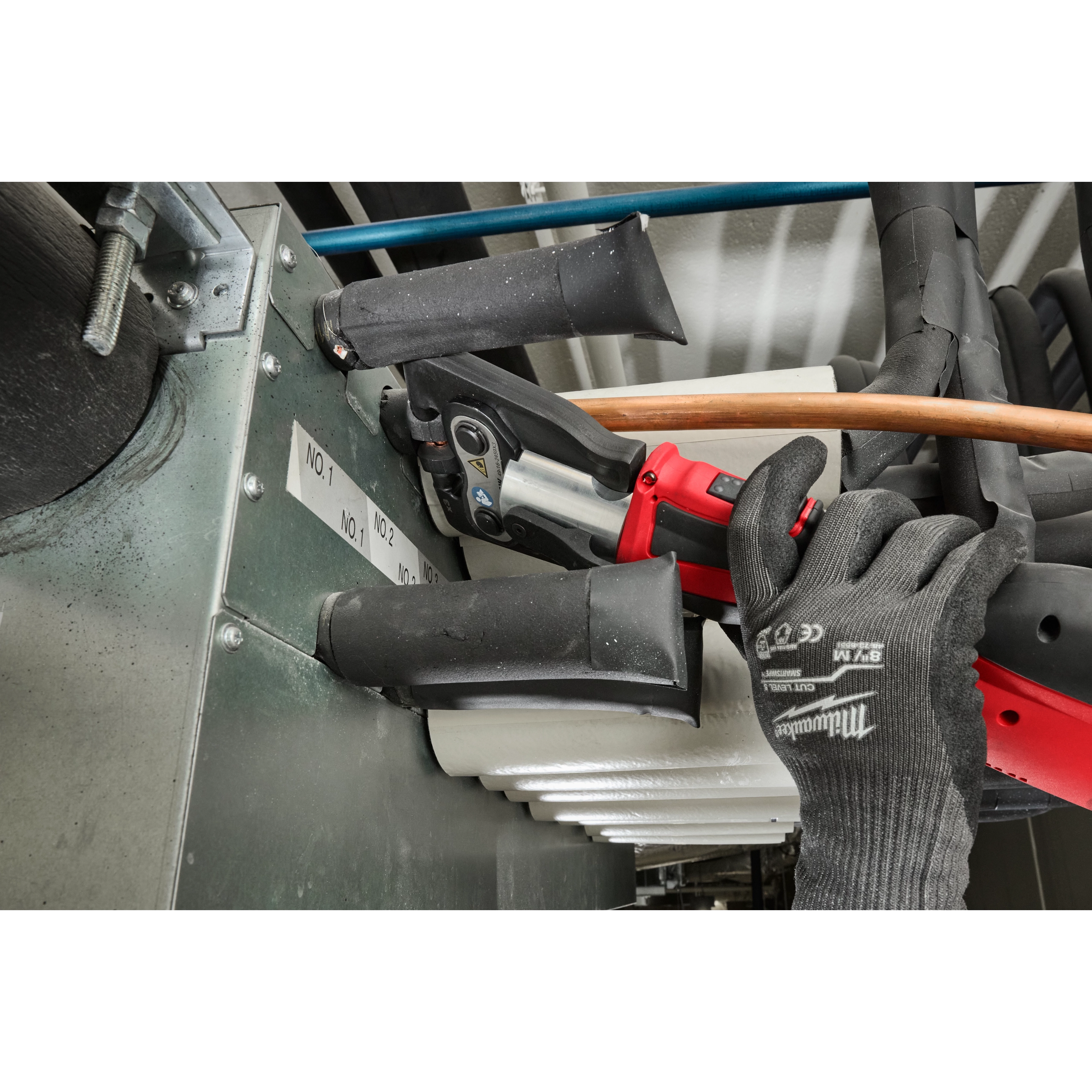 The ZoomLock MAX® & MaxiPro® Pivoting Press Ring Kit for M18™ FORCE LOGIC™ Press Tool is being used in a mechanical setting. A gloved hand operates the red press tool, handling pipes connected to a metal surface with labeled tags and several black and blue features for pipe fittings visible.