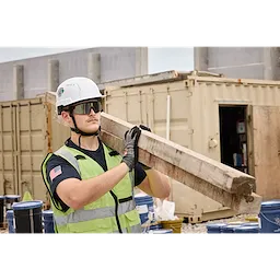 A construction worker in a safety vest and helmet carries a wooden plank on his shoulder. He wears Wrap Around Safety Glasses - Mirrored Anti-Scratch Lenses and black gloves at a busy construction site with containers and barrels in the background.