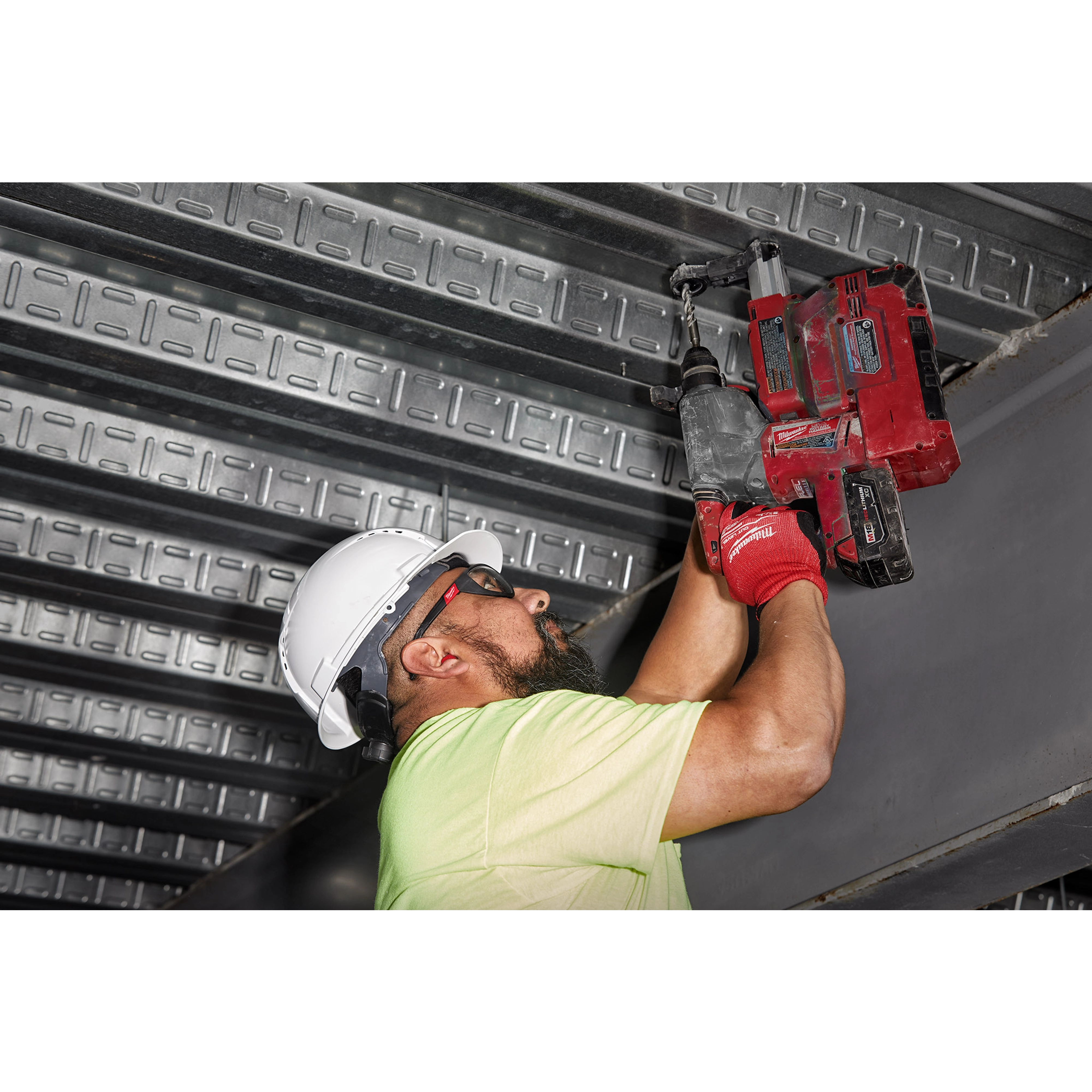 Worker wearing a hard hat and safety gloves uses a red power drill on a metal structure. Ear plugs are recommended for noise protection.