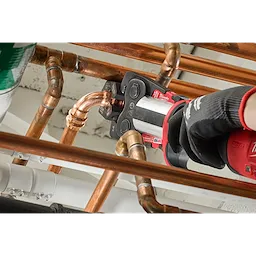 A gloved hand operates an M18™ FORCE LOGIC™ Press Tool to secure a copper pipe fitting using the 1/4" - 7/8" Streamline® ACR Pivoting Press Ring Kit. The tool is photographed amidst a network of copper pipes, demonstrating the pivoting press ring’s function in a plumbing installation.
