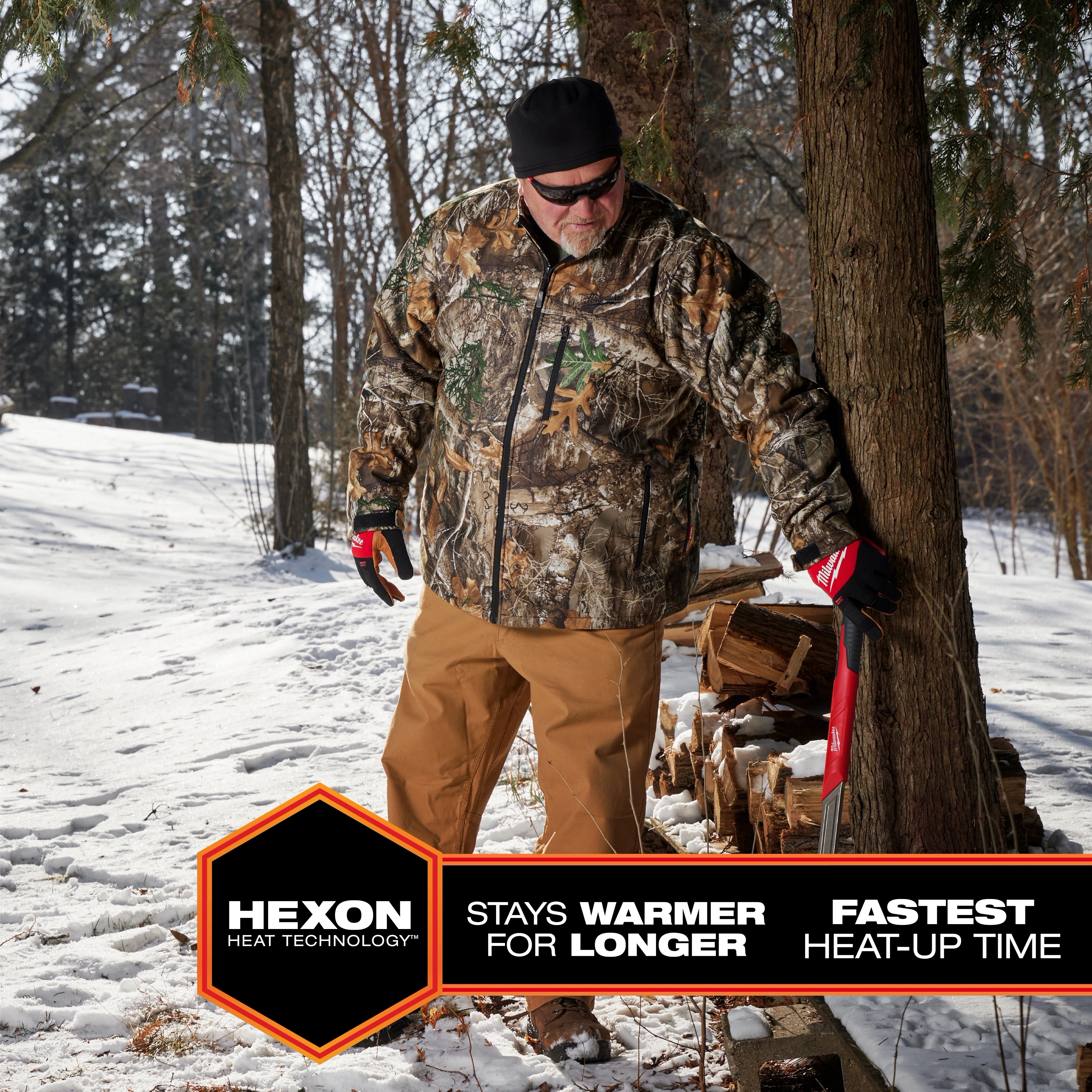 Person wearing M12 Heated QUIETSHELL Jacket in camo pattern, standing outdoors in a snowy forest, carrying an axe by a woodpile.