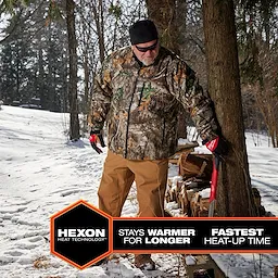 Person wearing M12 Heated QUIETSHELL Jacket in camo pattern, standing outdoors in a snowy forest, carrying an axe by a woodpile.