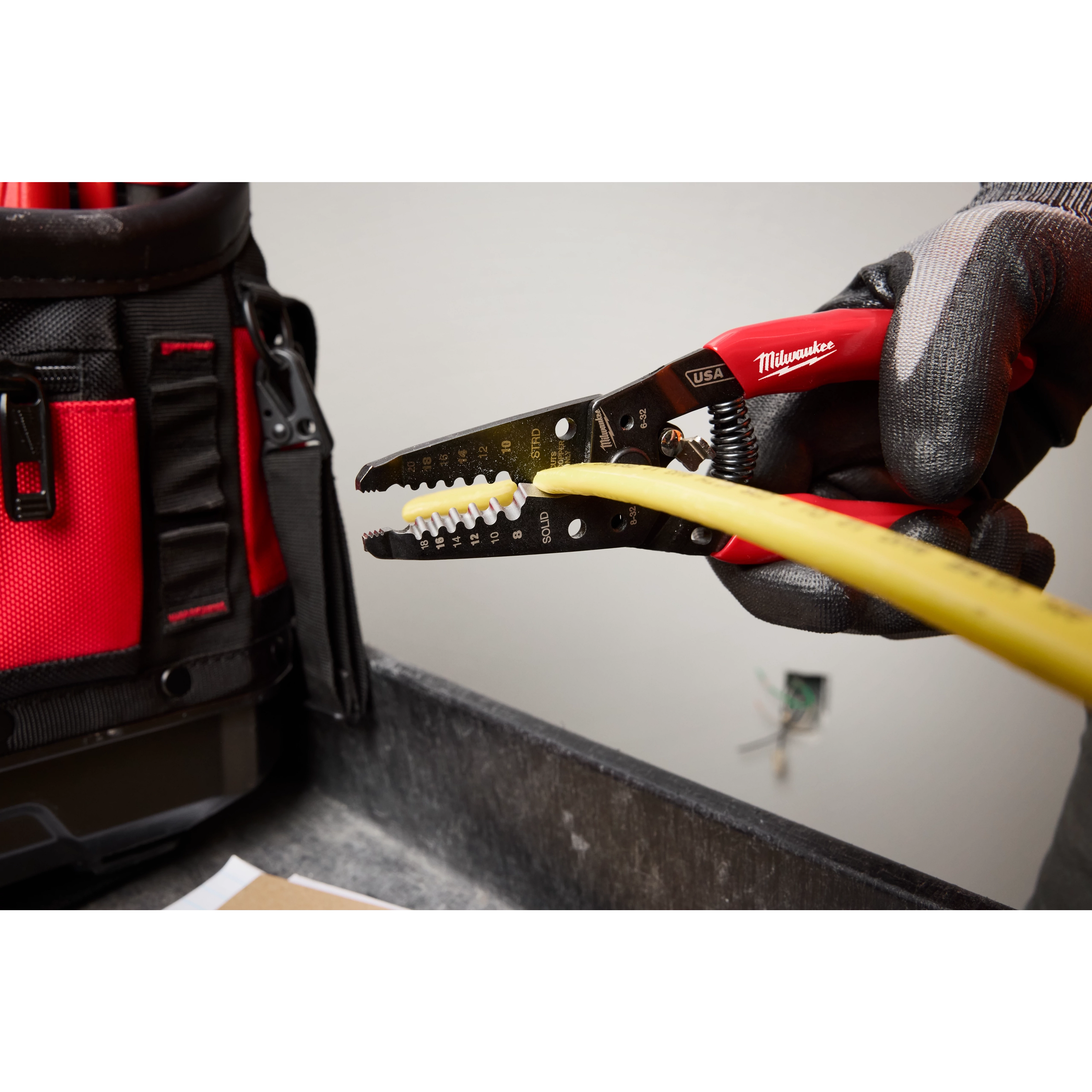 Gloved hand using the 8-20 AWG Dipped Grip Wire Stripper & Cutter (USA) to strip the insulation from a yellow wire, with a tool bag nearby.