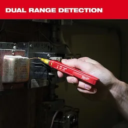 A person's hand holds a Milwaukee dual-range detection tool against an industrial electrical panel to test voltage. The bright red tool is labeled with voltage ranges and features an illuminated tip for precise work. The image emphasizes the tool's functionality in a technical environment.