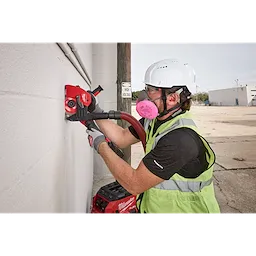 A worker uses a power tool equipped with the Milwaukee 6" Tuckpointing Dust Shroud to work on a concrete wall. The dust shroud is attached to a vacuum system, minimizing dust during the task.