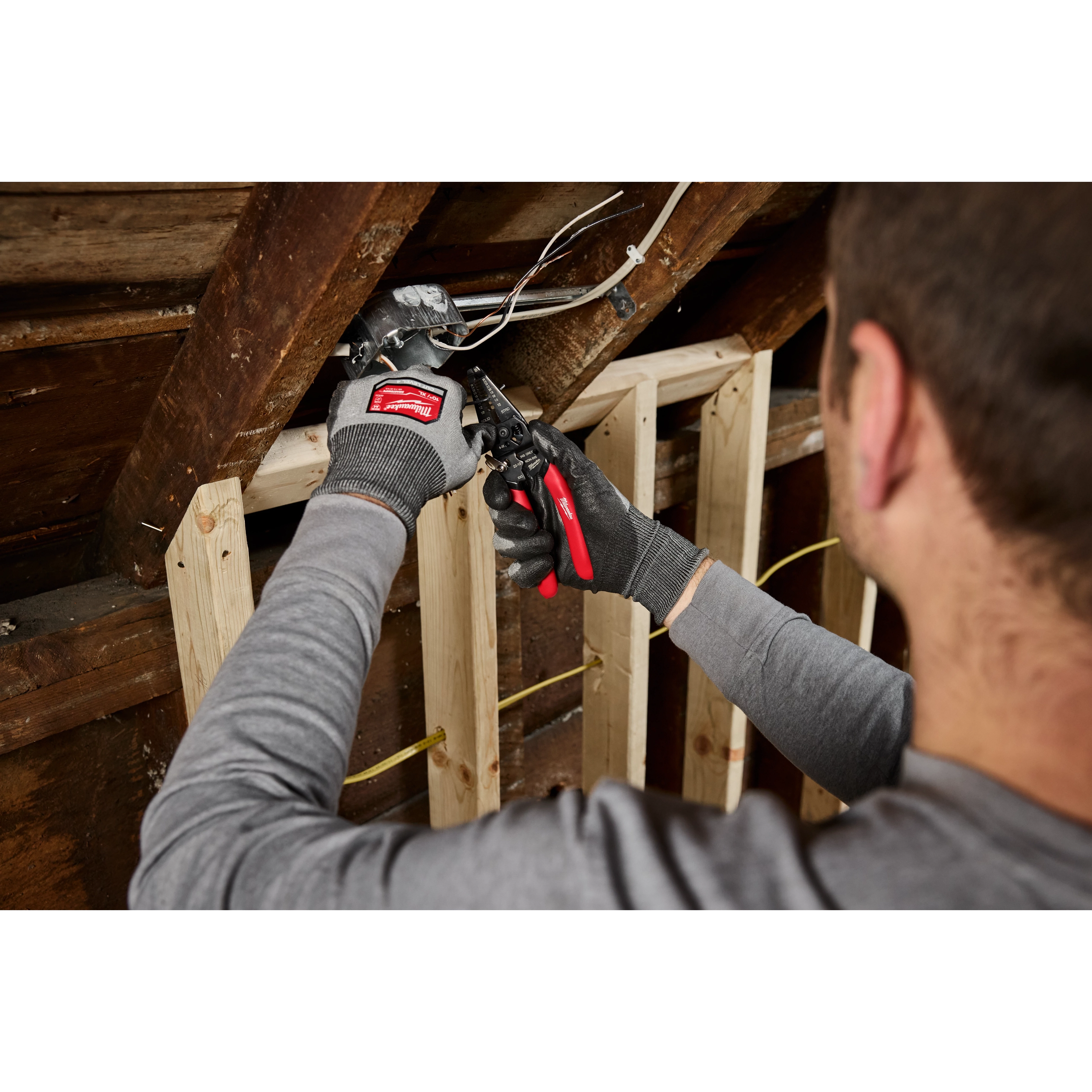 A person wearing gloves uses a 10-28 AWG Multi-Purpose Dipped Grip Wire Stripper & Cutter w/ Reinforced Head on an electrical box in an attic. The tool has red and black handles and is being used to strip wires.
