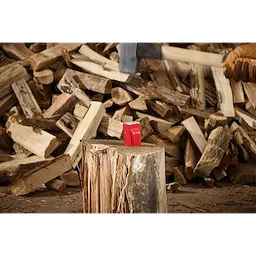 The 5lb Splitting Wedge is placed on a large log ready for splitting, surrounded by stacks of chopped firewood. A hand wearing a work glove holds an axe poised above the wedge, about to strike. The scene is set in a woodworking area.