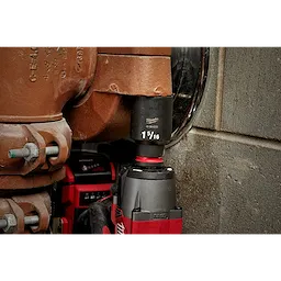 The SHOCKWAVE%20Impact%20Duty%E2%84%A2%201%2F2%22%20Drive%201-5%2F16%22%20Deep%206%20Point%20Socket is attached to a power tool, being utilized to tighten a large bolt on an industrial pipe fitting. The socket is black and labeled with size 1-5/16".