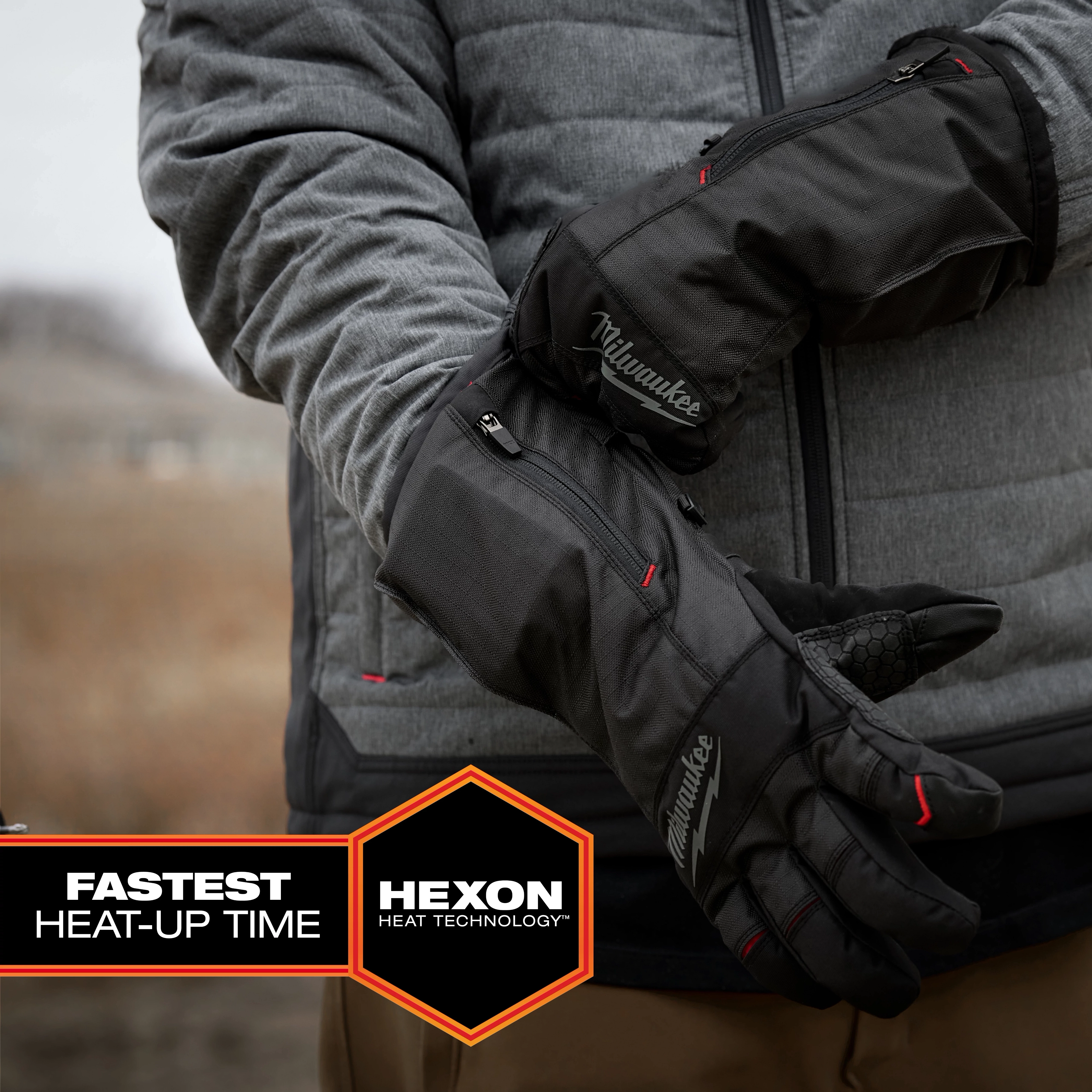 Person wearing black Milwaukee Heated Winter Gloves with zippers, standing outdoors in a gray jacket. The gloves feature Hexon heat technology and have the fastest heat-up time, as indicated by the text in the bottom left corner. Background includes blurred landscape and soft lighting.