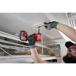 A person uses the M18 FUEL™ SURGE™ 1/4" Hex Hydraulic Driver to secure a metal bracket to a ceiling. The tool is red and black, and the user is wearing grey gloves with green trim. Various pipes and cables are visible in the background.
