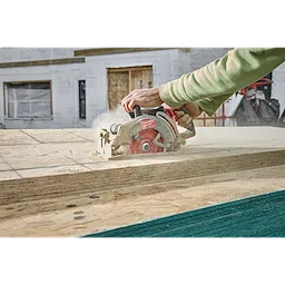 A person operates a M18 FUEL™ 7-1/4" Rear Handle Circular Saw to cut through wooden boards at a construction site. Dust is visible around the cutting area, indicating active use. A partially constructed building is in the background.