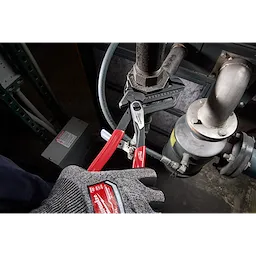 A gloved hand is using 12" Pliers Wrench with red handles to tighten a pipe fitting. The wrench has textured jaws for gripping.