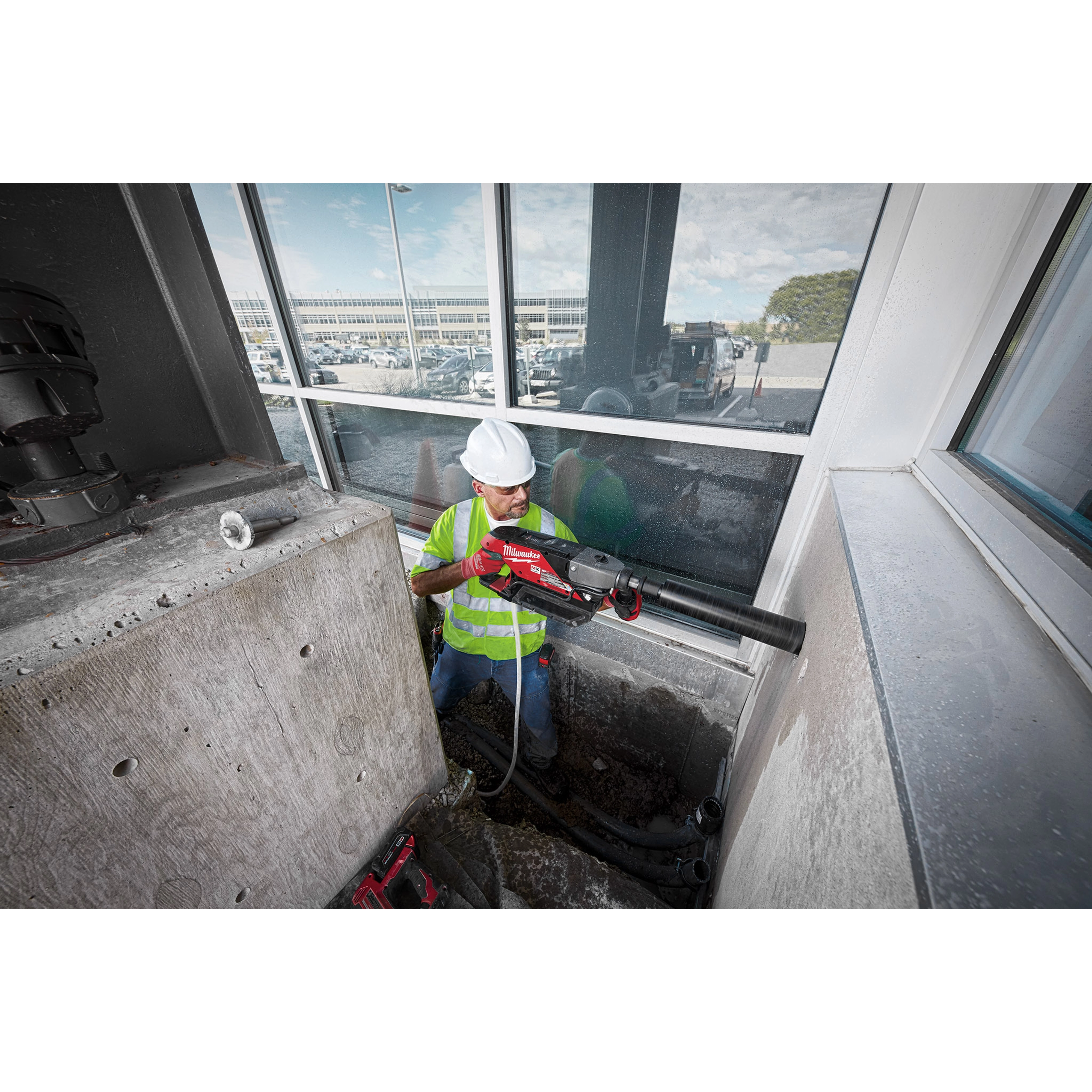 Worker using MX FUEL Handheld Core Drill to cut through concrete in an industrial worksite, wearing safety gear and high-visibility vest.