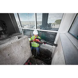 Worker using MX FUEL Handheld Core Drill to cut through concrete in an industrial worksite, wearing safety gear and high-visibility vest.