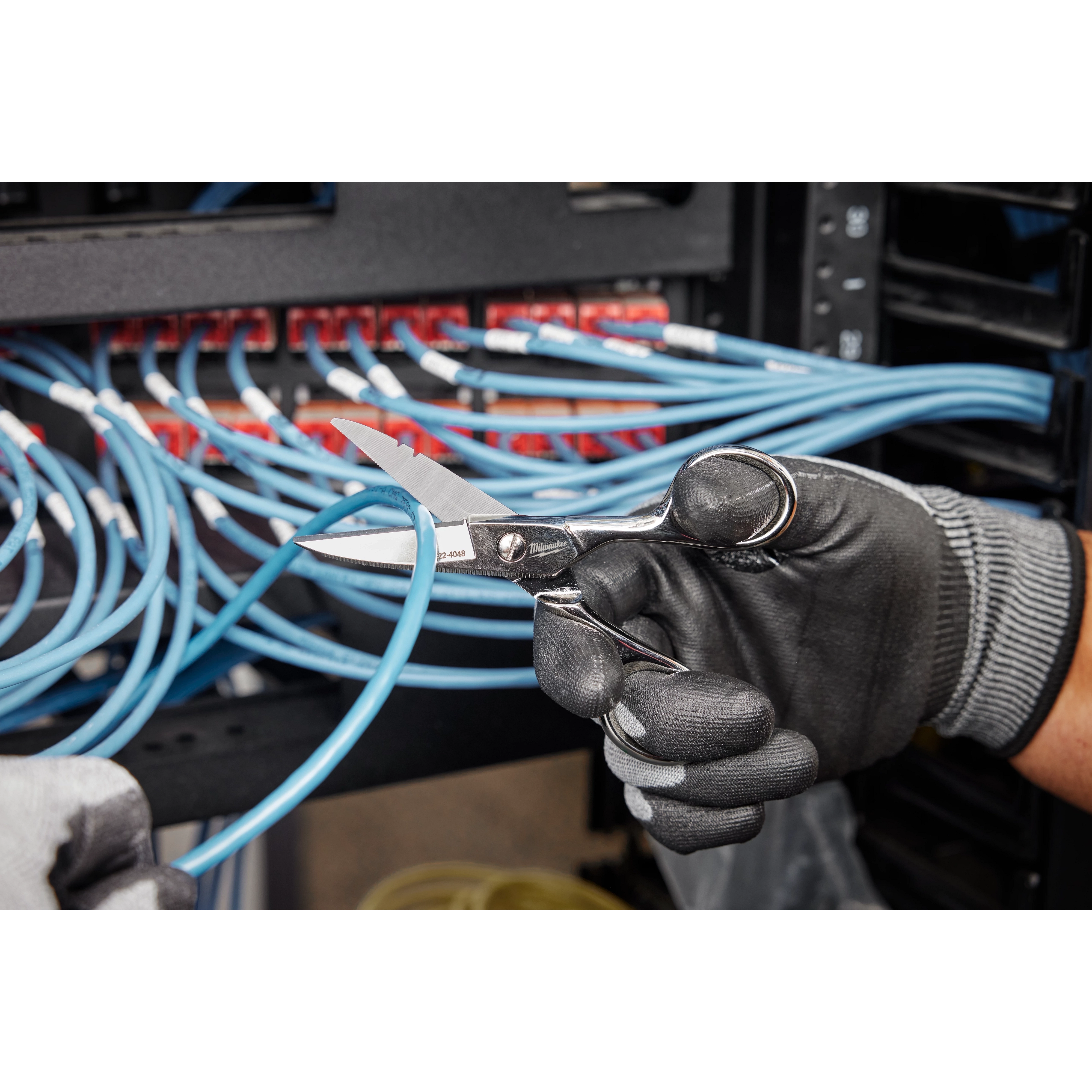 A gloved hand holds Electrician Scissors, cutting through blue electrical wires amidst a network panel.