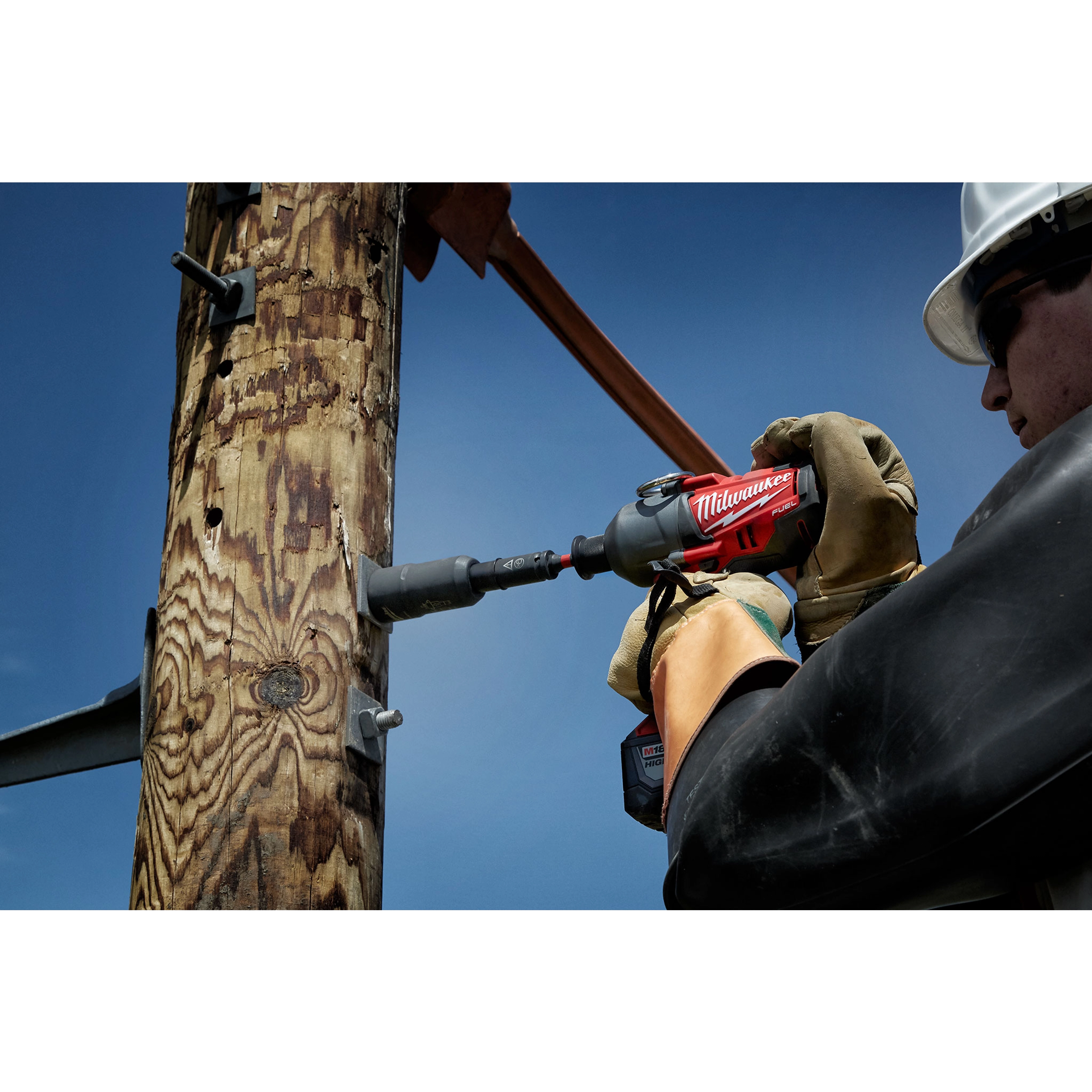 Worker uses a red Power Utility Socket to drill a hole in a wooden utility pole against a clear blue sky background.
