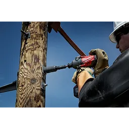 Worker uses a red Power Utility Socket to drill a hole in a wooden utility pole against a clear blue sky background.