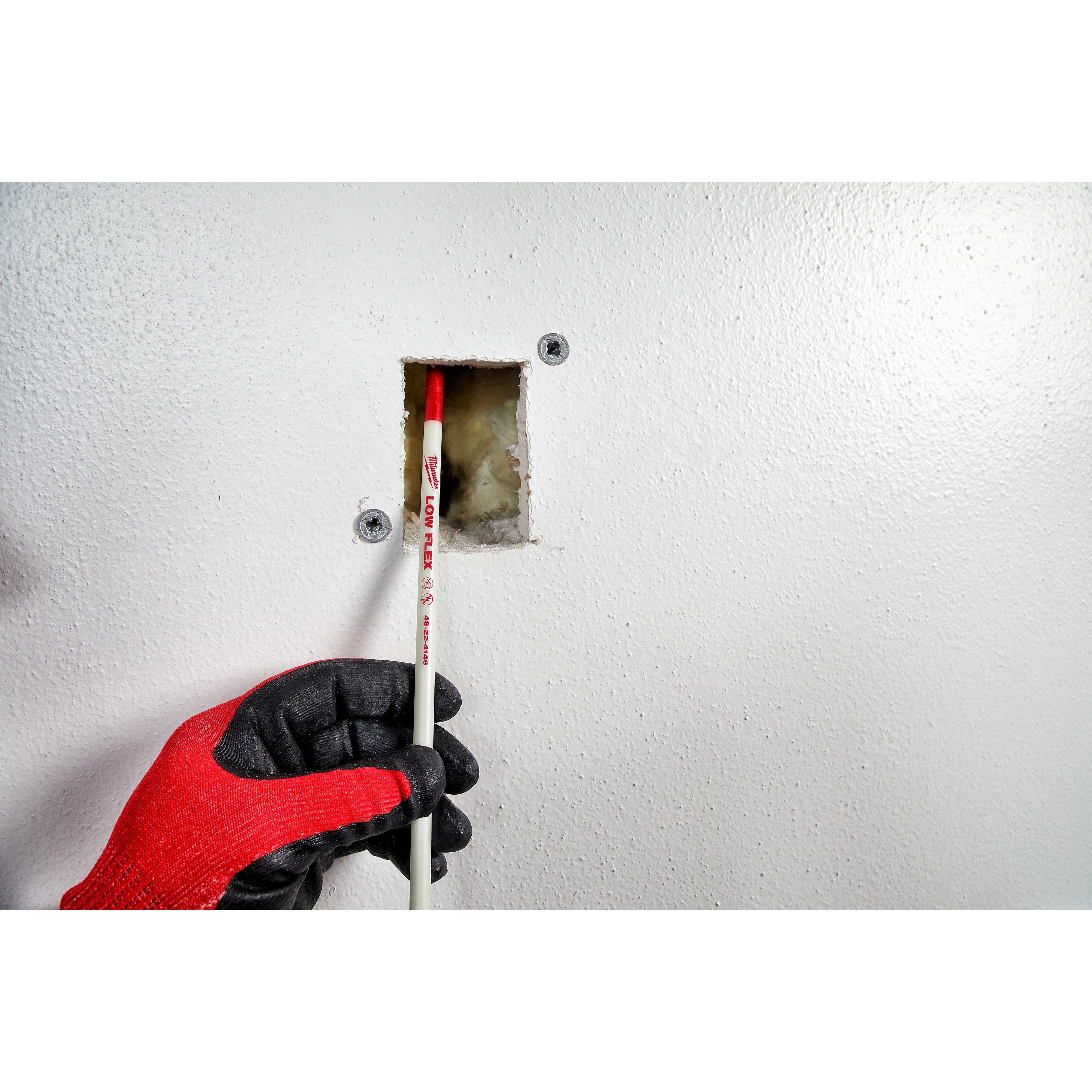 A worker uses the 5' Low Flex Fish Stick to guide wiring through a small wall opening while wearing black and red gloves.