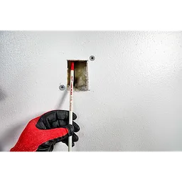 A worker uses the 5' Low Flex Fish Stick to guide wiring through a small wall opening while wearing black and red gloves.