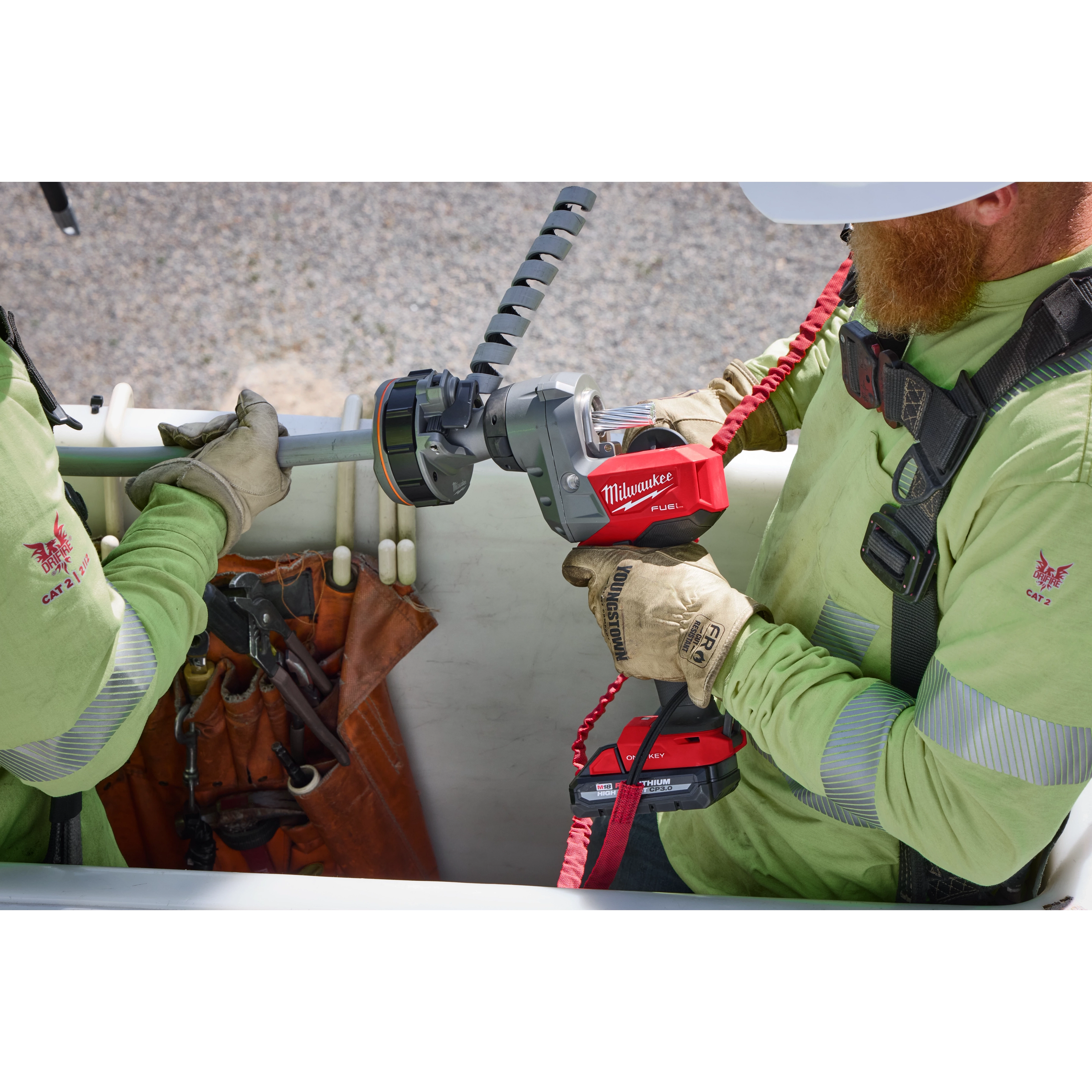 Two workers use the M18 FUEL™ Covered Conductor Cable Stripper to strip a cable. One worker holds the cable while the other operates the red and black handheld device. Both are wearing safety harnesses and green work shirts.