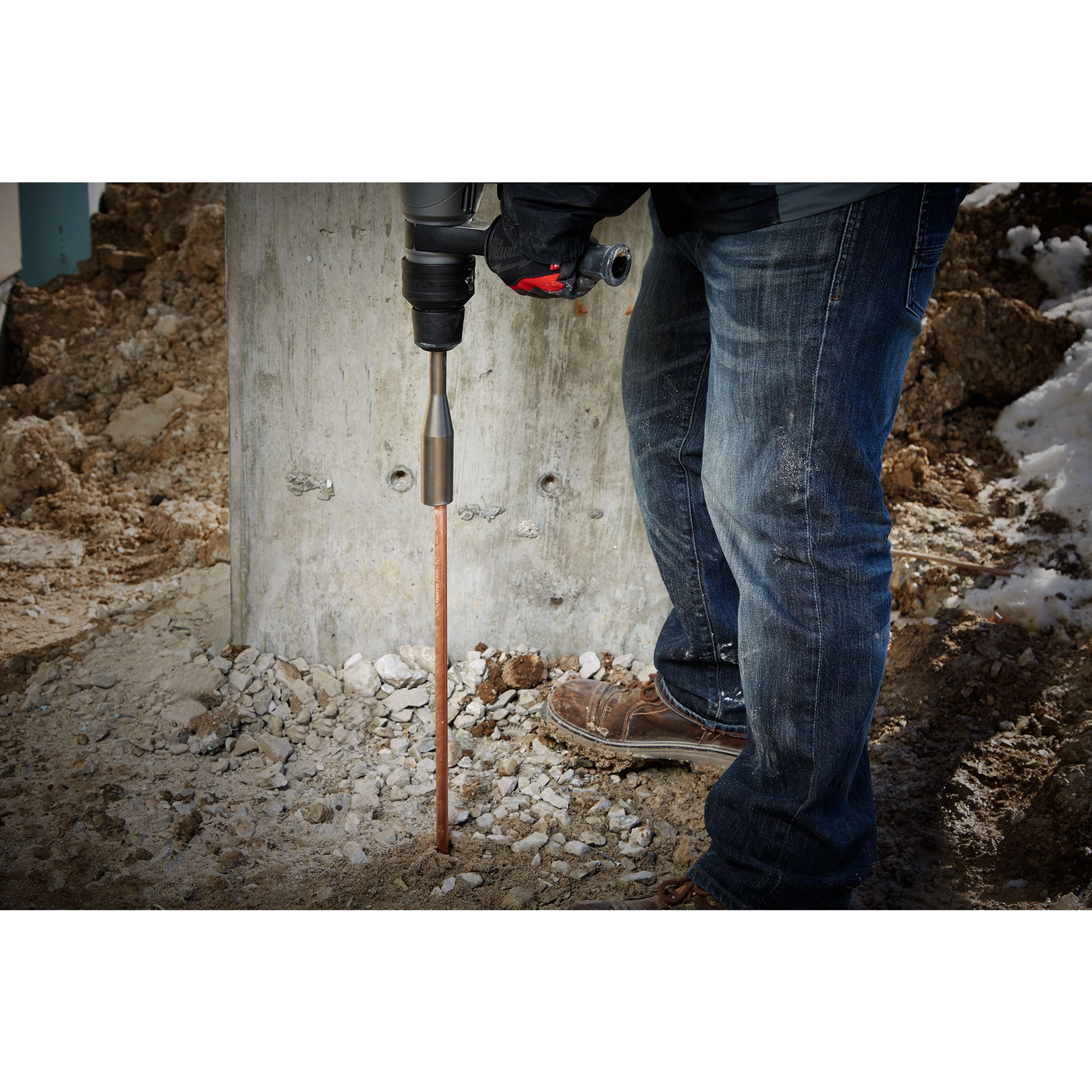 A worker wearing jeans uses an SDSMAX GROUND ROD DRIVER to install a copper ground rod into the rocky ground at a construction site.