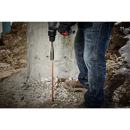 A worker wearing jeans uses an SDSMAX GROUND ROD DRIVER to install a copper ground rod into the rocky ground at a construction site.