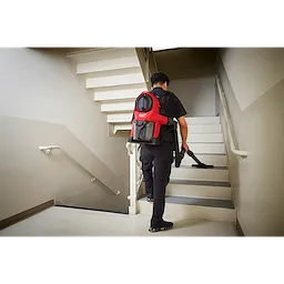 Person carrying and using the M18 FUEL™ 3-in-1 Backpack Vacuum while walking up a stairwell. The vacuum is red with black straps and has multiple compartments for different cleaning functions.