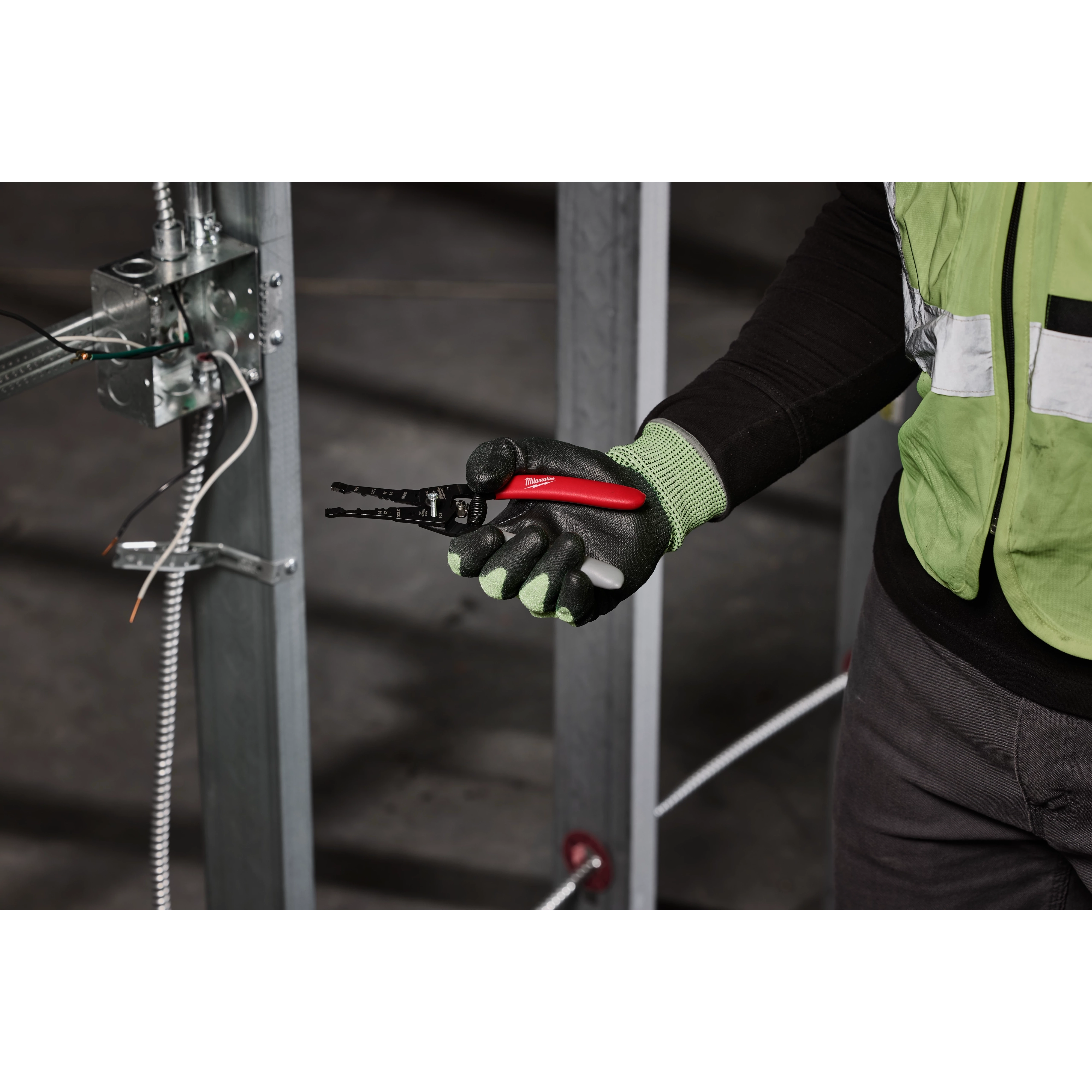 A person in a safety vest and gloves holds a 12/2 & 14/2 NM Dipped Grip Wire Stripper & Cutter. The tool has a black and red handle and is shown in a construction environment with exposed wiring.