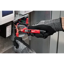 Person wearing gloves uses a Dual Range Voltage Detector w/ LED on a bundle of wires inside an electrical box. The tool's LED lights up, indicating voltage.