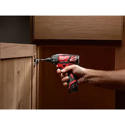 A person using the red and black cordless M12 Screwdriver to fasten a hinge on a wooden cabinet door.