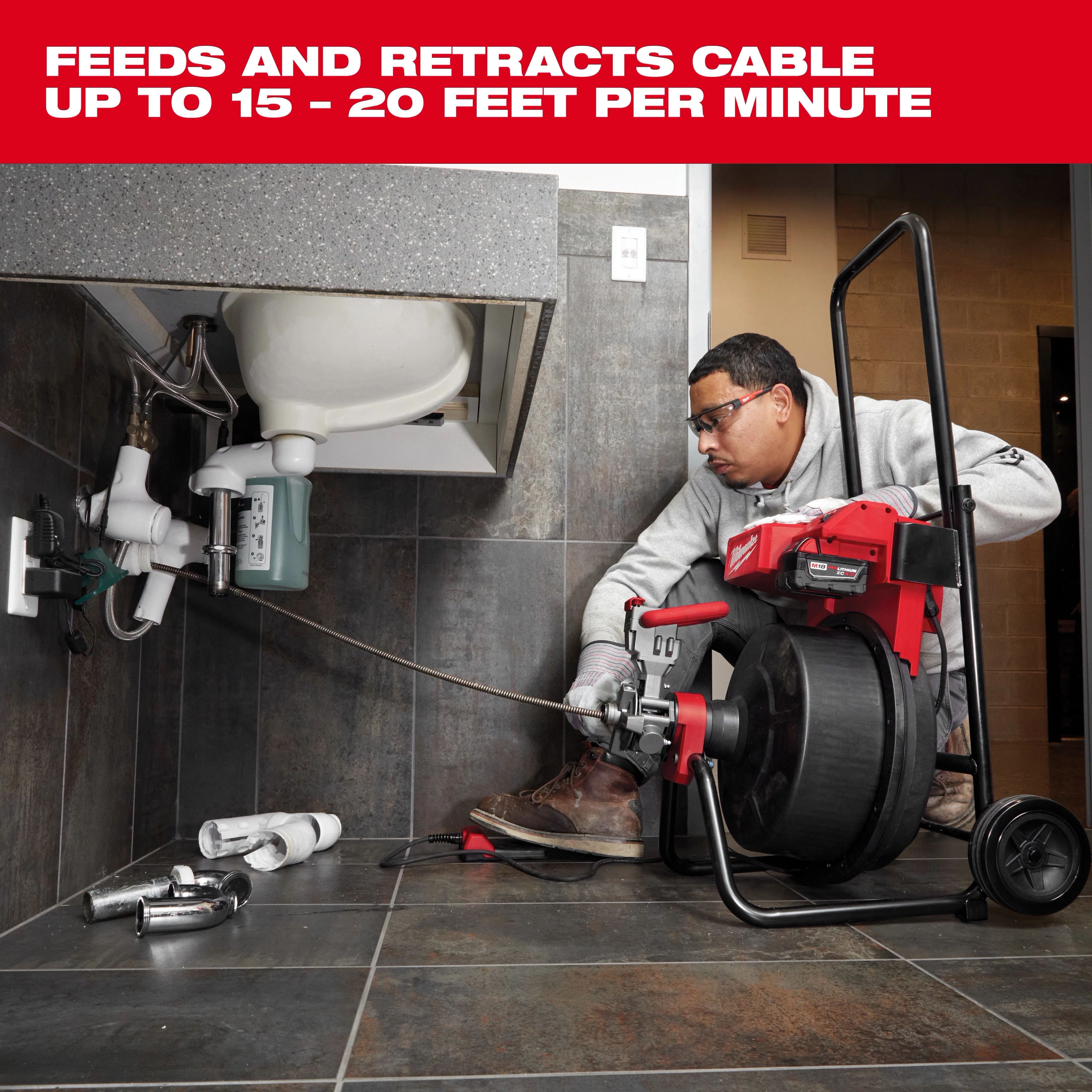 Man using the Milwaukee drain cleaner under a sink. The tool can feed and retract cables at speeds of 15-20 feet per minute. The workspace includes plumbing tools and components on a tiled floor.