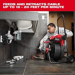 Man using the Milwaukee drain cleaner under a sink. The tool can feed and retract cables at speeds of 15-20 feet per minute. The workspace includes plumbing tools and components on a tiled floor.