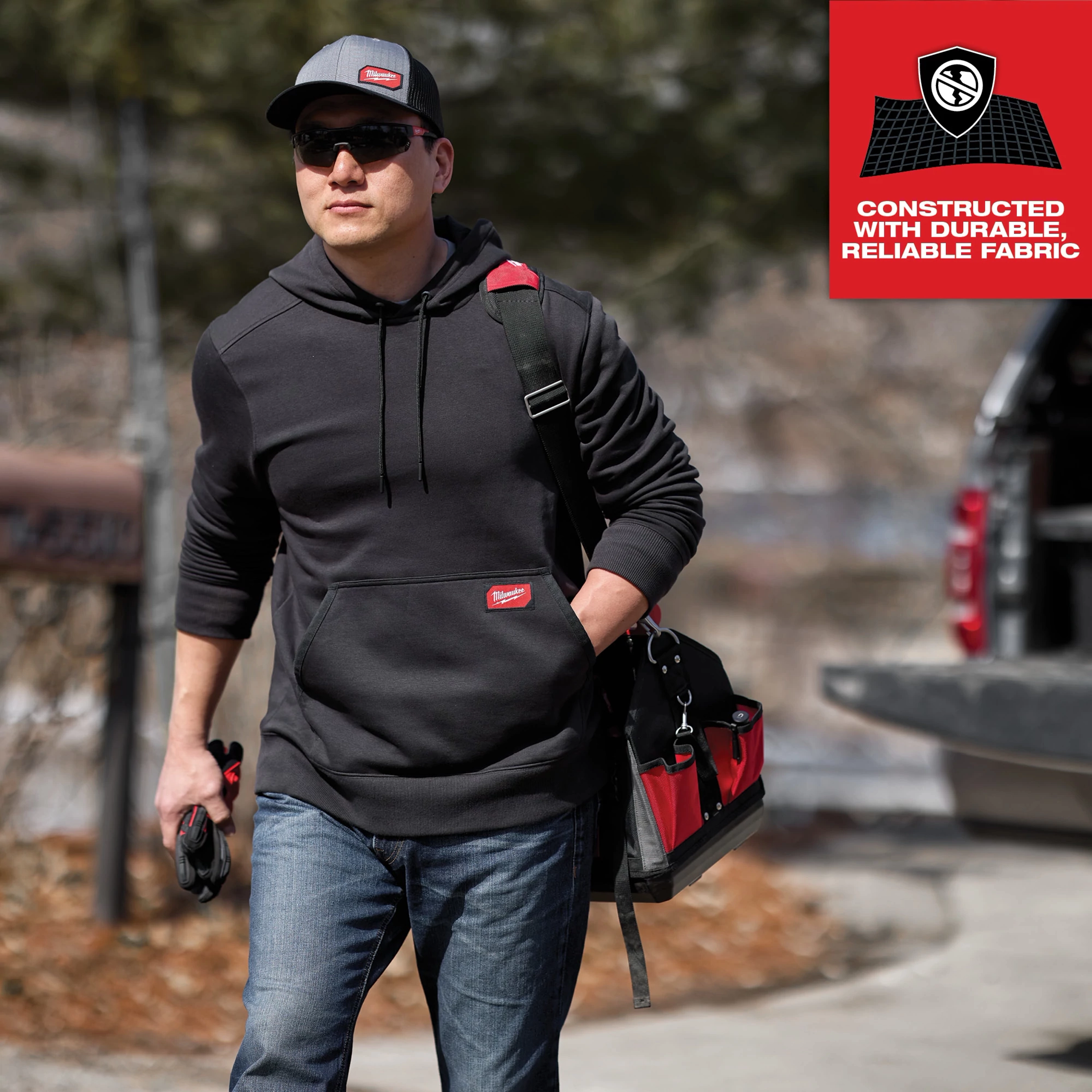 A person wearing a dark gray hoodie and blue jeans carries a tool bag while wearing a GridIron Snapback Trucker Hat - Gray.