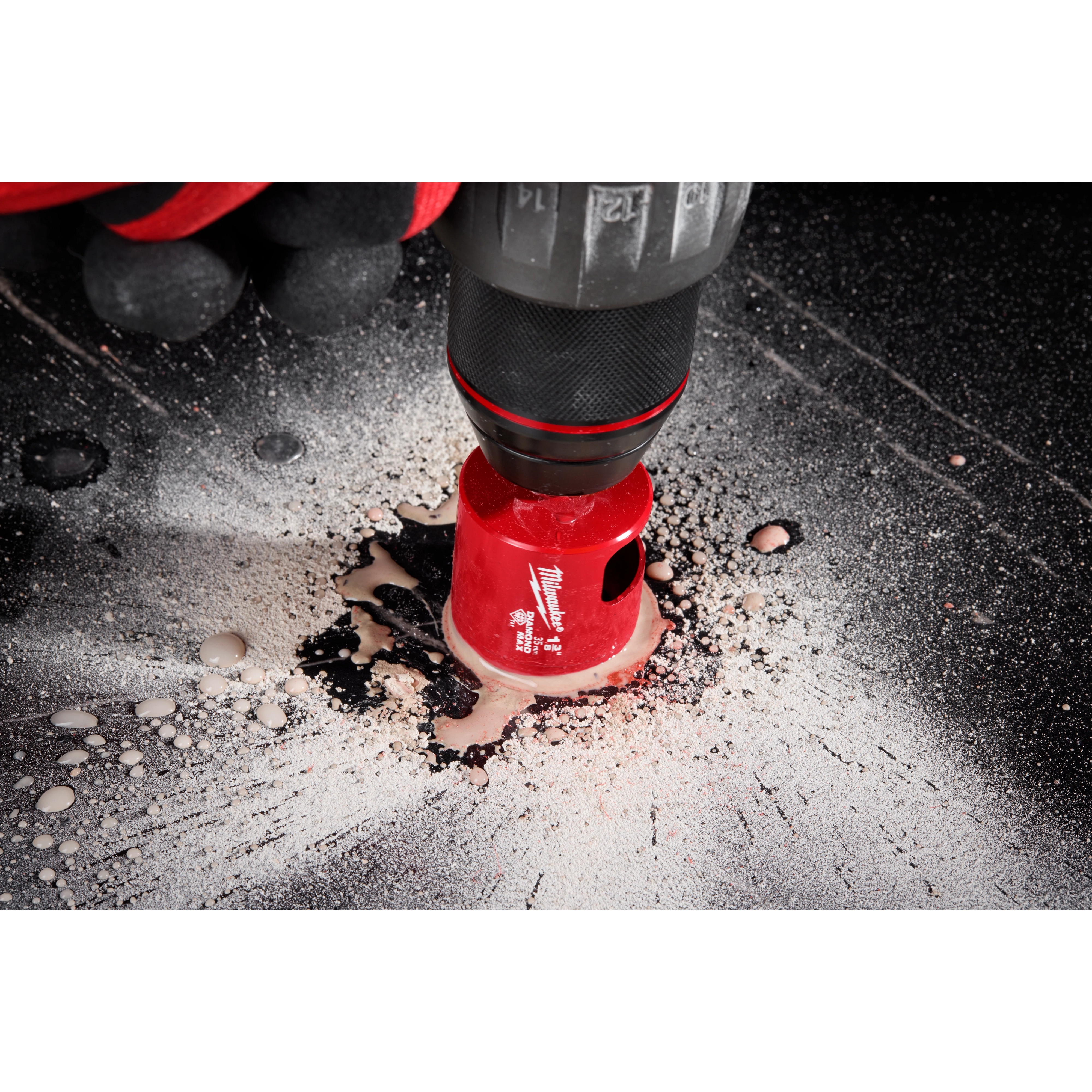 A close-up of a Milwaukee® 1-3/8" DIAMOND MAX™ Hole Saw Bit in use, cutting through a dark surface. The hole saw bit is red and attached to a drill, with debris and dust visible around the cutting area.