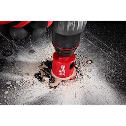 A close-up of a Milwaukee® 1-3/8" DIAMOND MAX™ Hole Saw Bit in use, cutting through a dark surface. The hole saw bit is red and attached to a drill, with debris and dust visible around the cutting area.