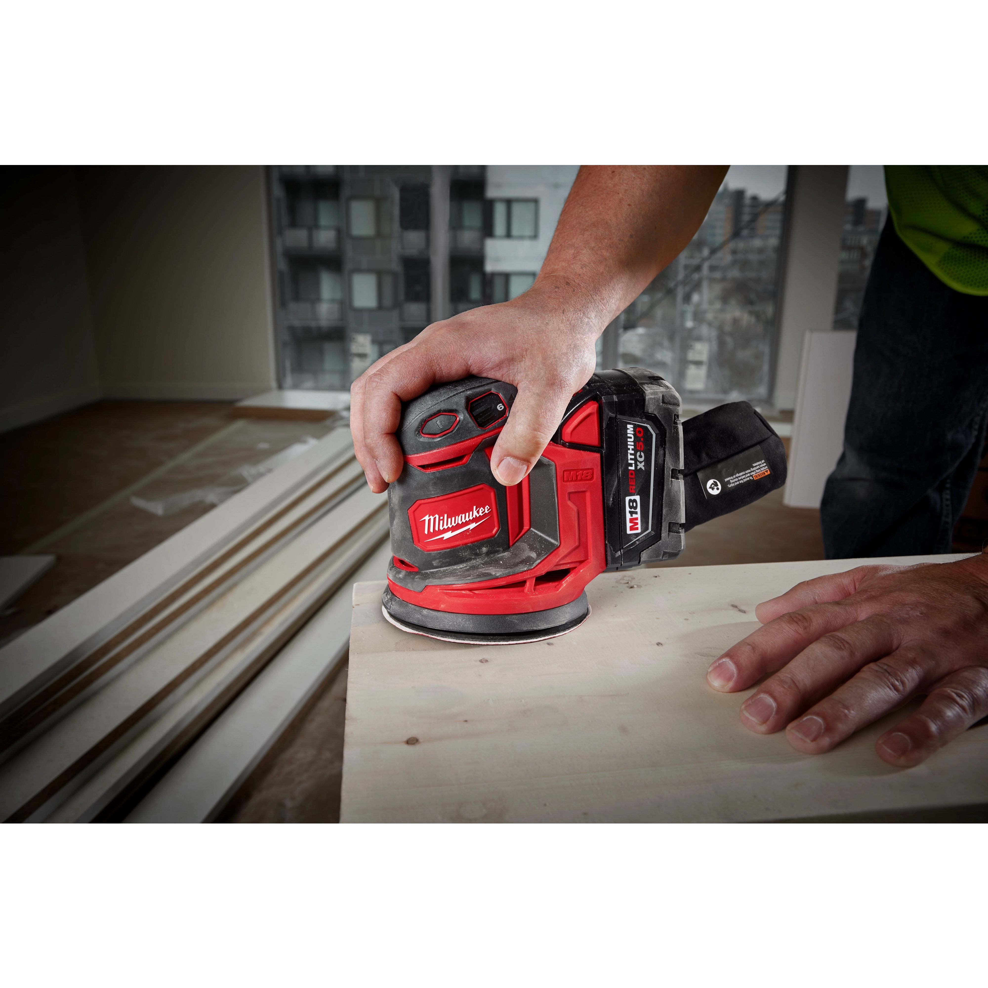 A person uses the M18 Random Orbit Sander to smooth a wooden surface. The sander has a red and black design and is cordless. The scene appears to be inside a room under construction, with visible building materials and high-rise buildings outside the window.