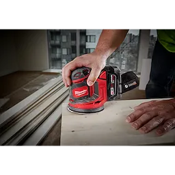 A person uses the M18 Random Orbit Sander to smooth a wooden surface. The sander has a red and black design and is cordless. The scene appears to be inside a room under construction, with visible building materials and high-rise buildings outside the window.