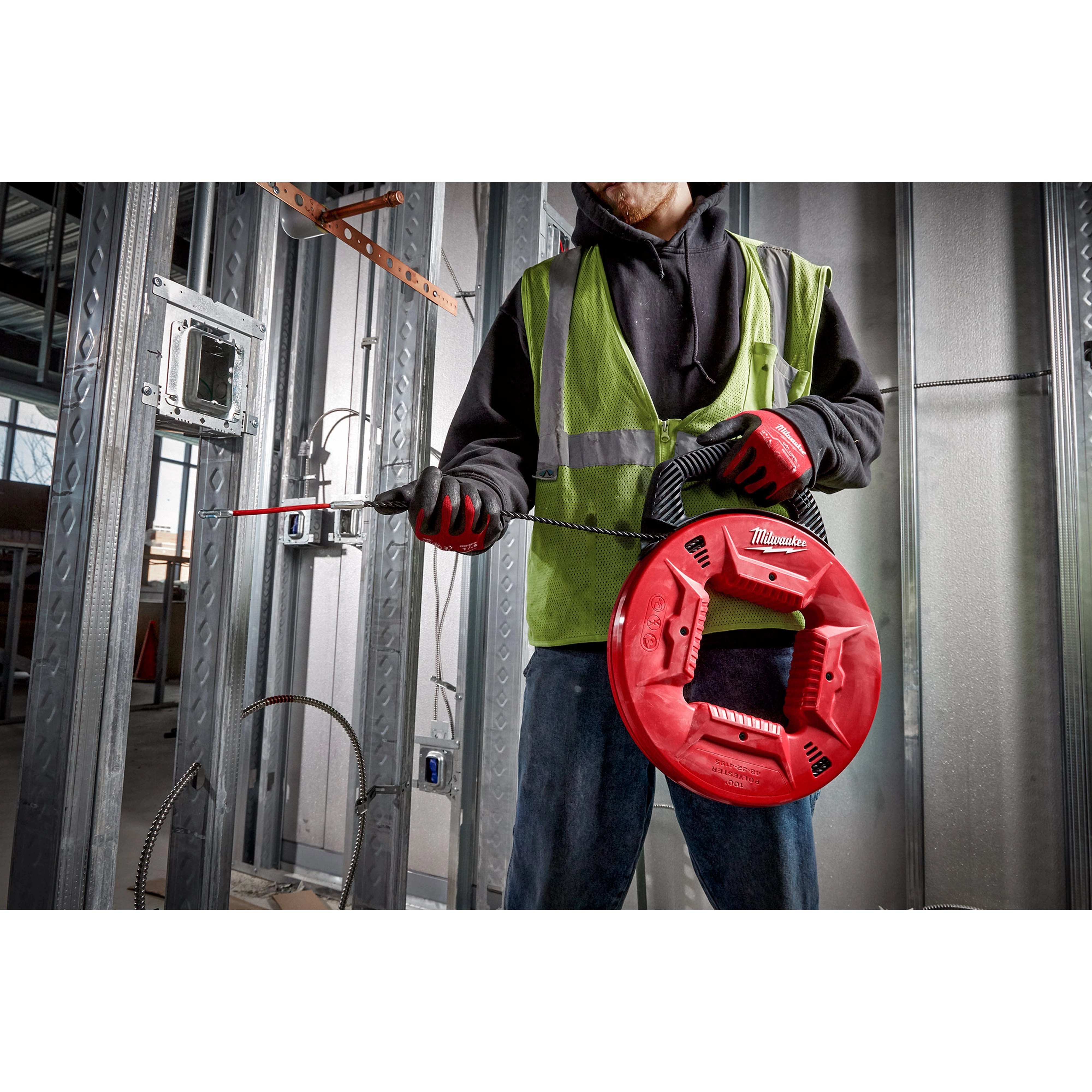Worker using a 100% Polyester Fish Tape with Flexible Leader in a metal-framed construction site, wearing a green safety vest and red gloves.