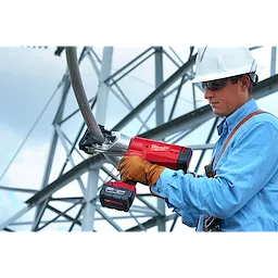 A worker wearing protective gear operates the FORCELOGIC™ M18™ 1590 ACSR Cutter Kit, cutting through a thick cable. The tool is red and black, battery-powered, and designed for efficient cable cutting, shown in an industrial setting with metal structures in the background.