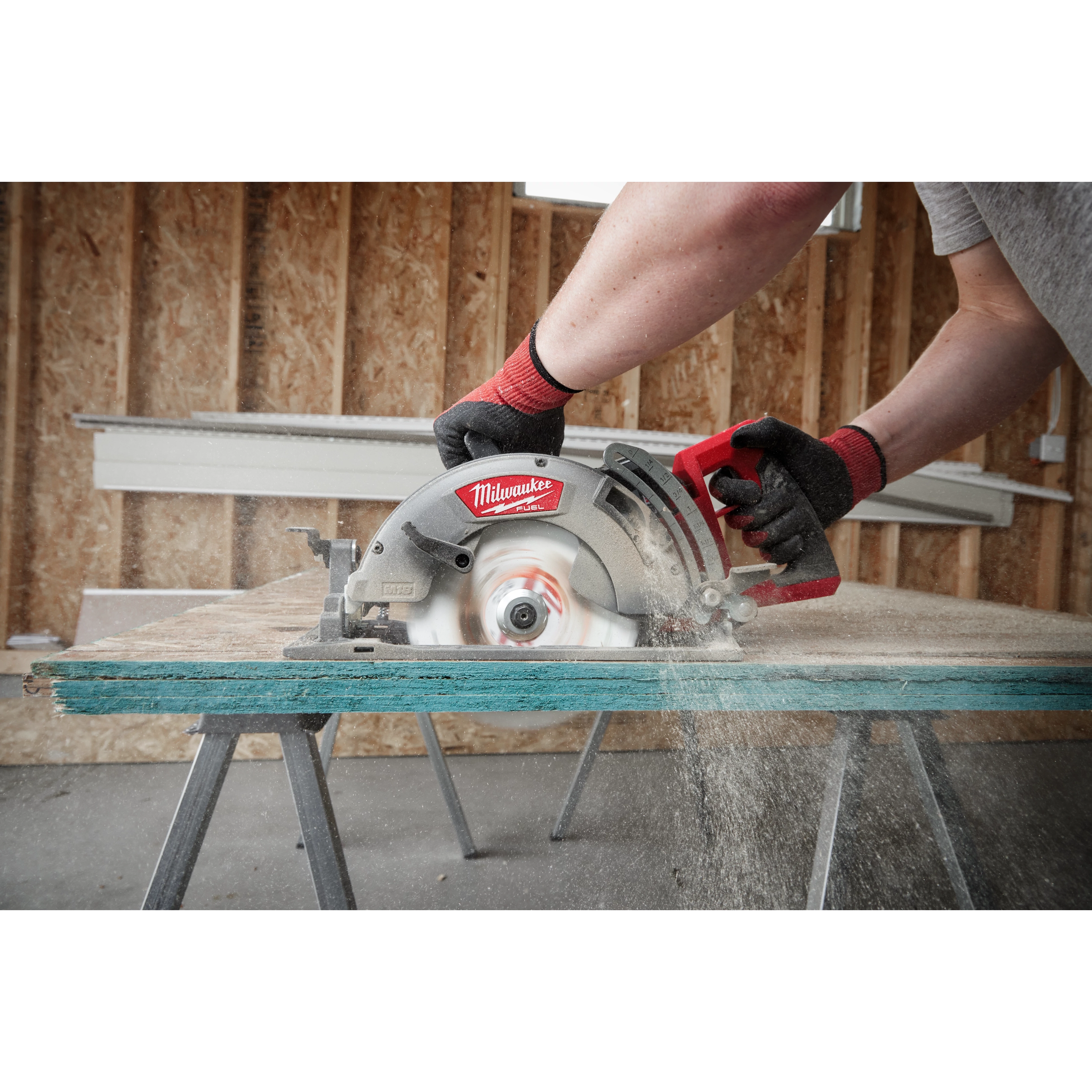 A person uses the M18 FUEL™ Rear Handle Circular Saw to cut through a piece of wood atop saw horses in a partially constructed building. The tool is in motion, creating sawdust, and the person is wearing gloves for protection.