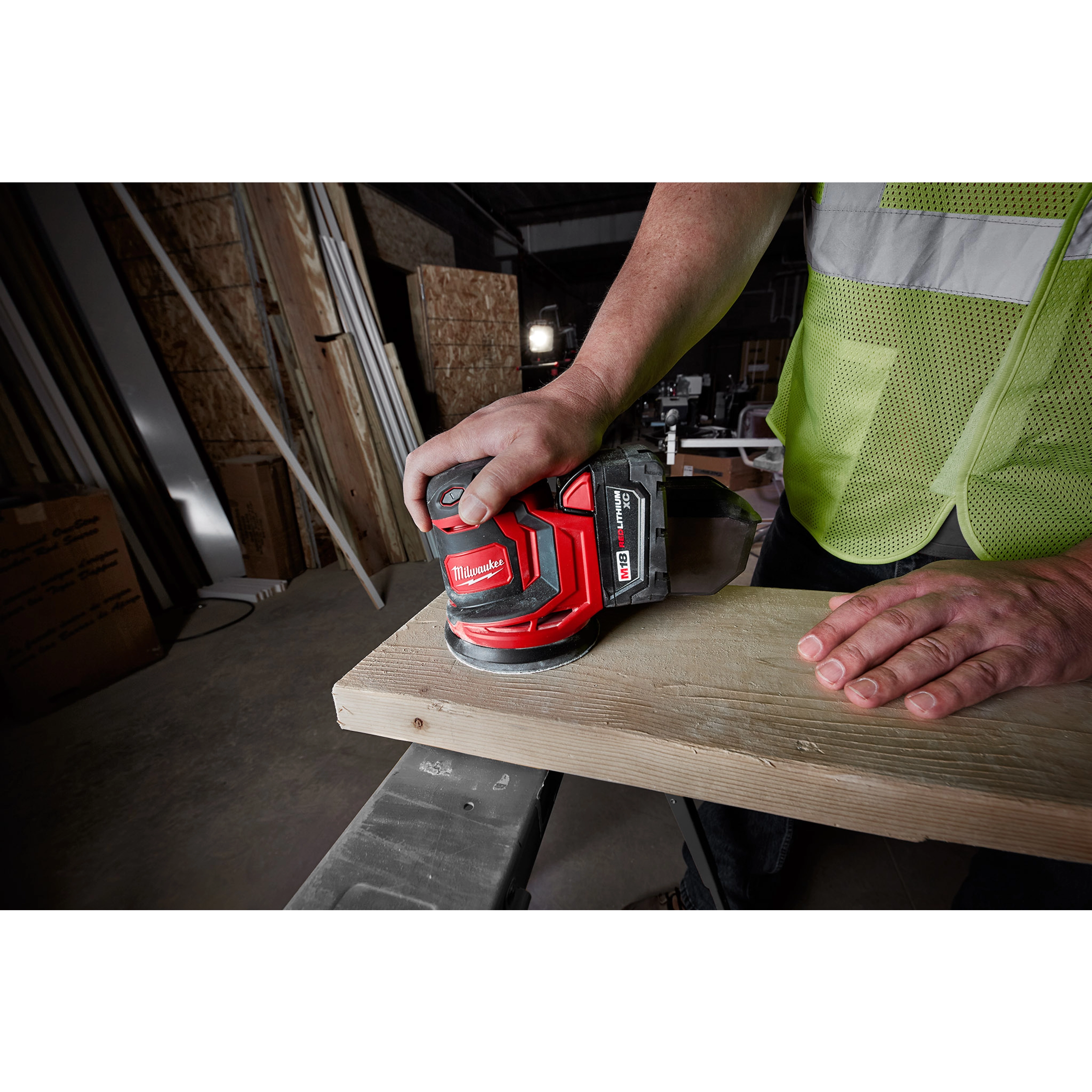 A person sands a wooden plank using an M18 Random Orbit Sander. The worker wears a reflective vest in a construction setting.