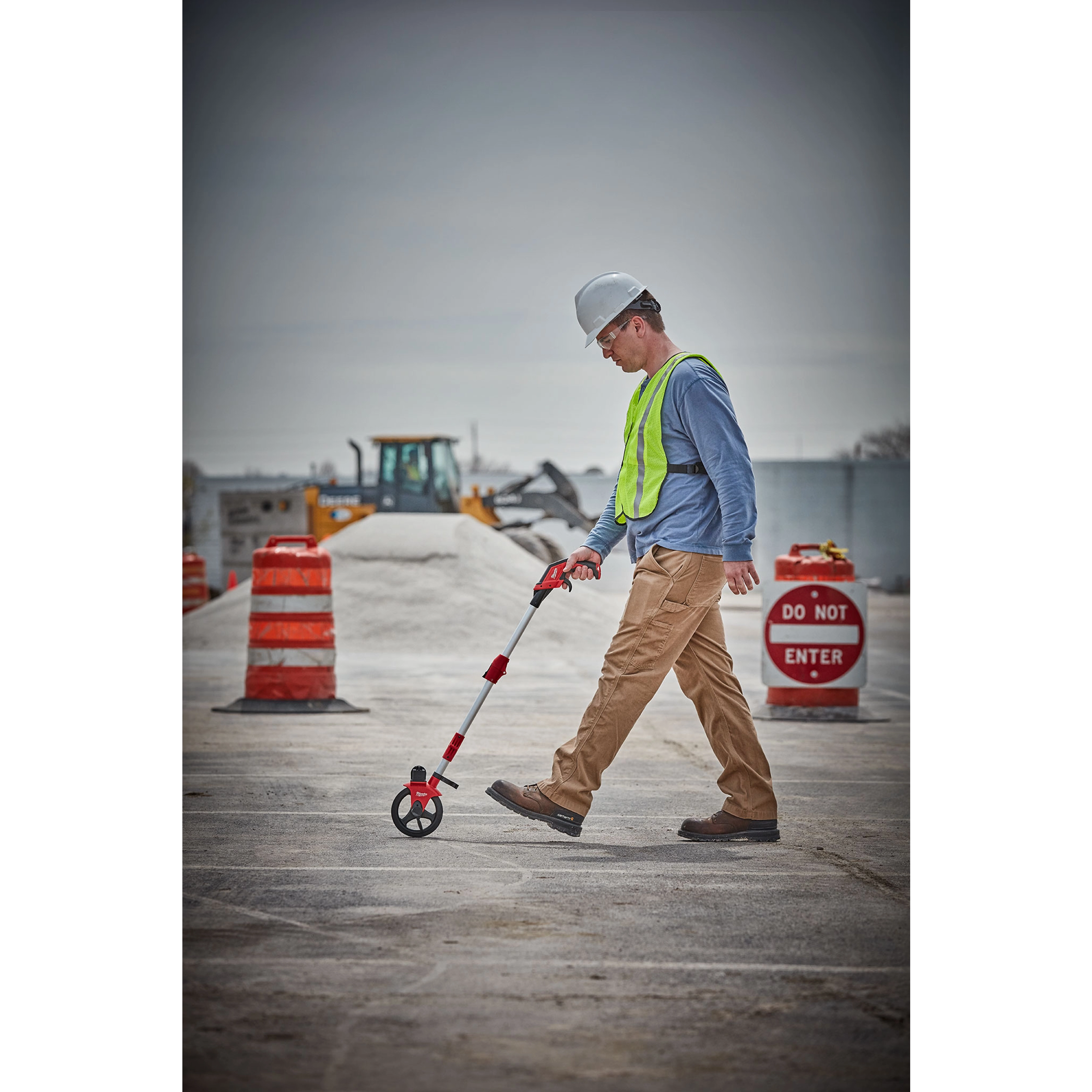 A construction worker uses a Measuring Wheel while walking on a concrete surface. Nearby, there are orange barrels and a "Do Not Enter" sign.