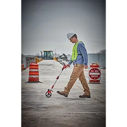 A construction worker uses a Measuring Wheel while walking on a concrete surface. Nearby, there are orange barrels and a "Do Not Enter" sign.