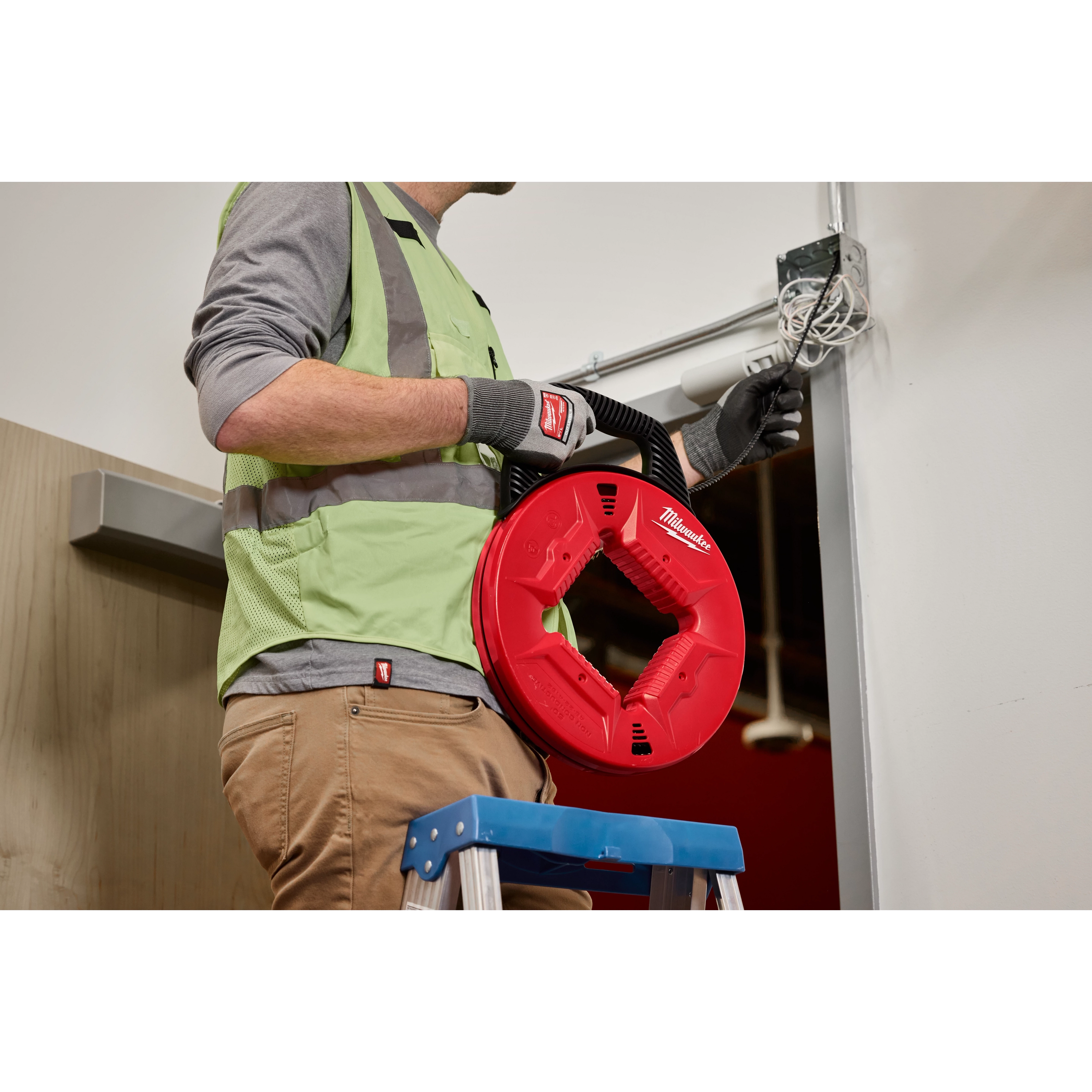 A person in workwear stands on a ladder feeding wire into a wall using 100’ Polyester Fish Tape with Nonconductive Tip. The fish tape is red and circular, branded with "Milwaukee".