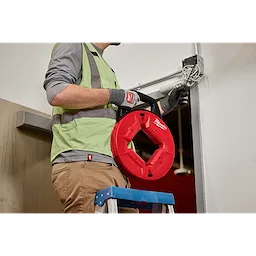 A person in workwear stands on a ladder feeding wire into a wall using 100’ Polyester Fish Tape with Nonconductive Tip. The fish tape is red and circular, branded with "Milwaukee".