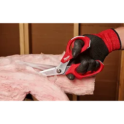 A hand wearing a black and red glove uses Offset Jobsite Scissors with red handles to cut pink insulation material. The background shows wooden framing and cardboard.