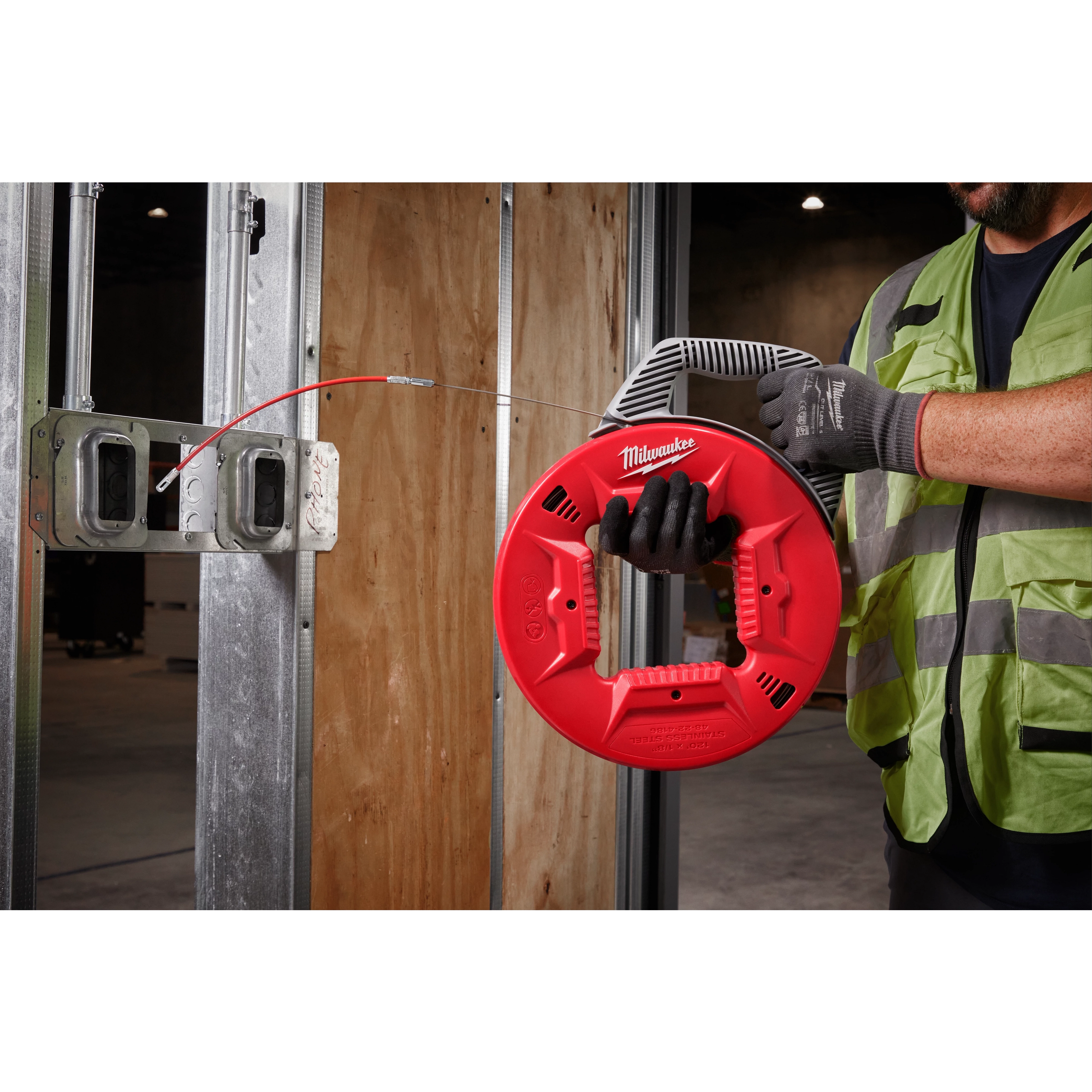 A construction worker uses a 120' 1/8" Stainless Steel Fish Tape with a red and black handle to thread wiring through metal studs.