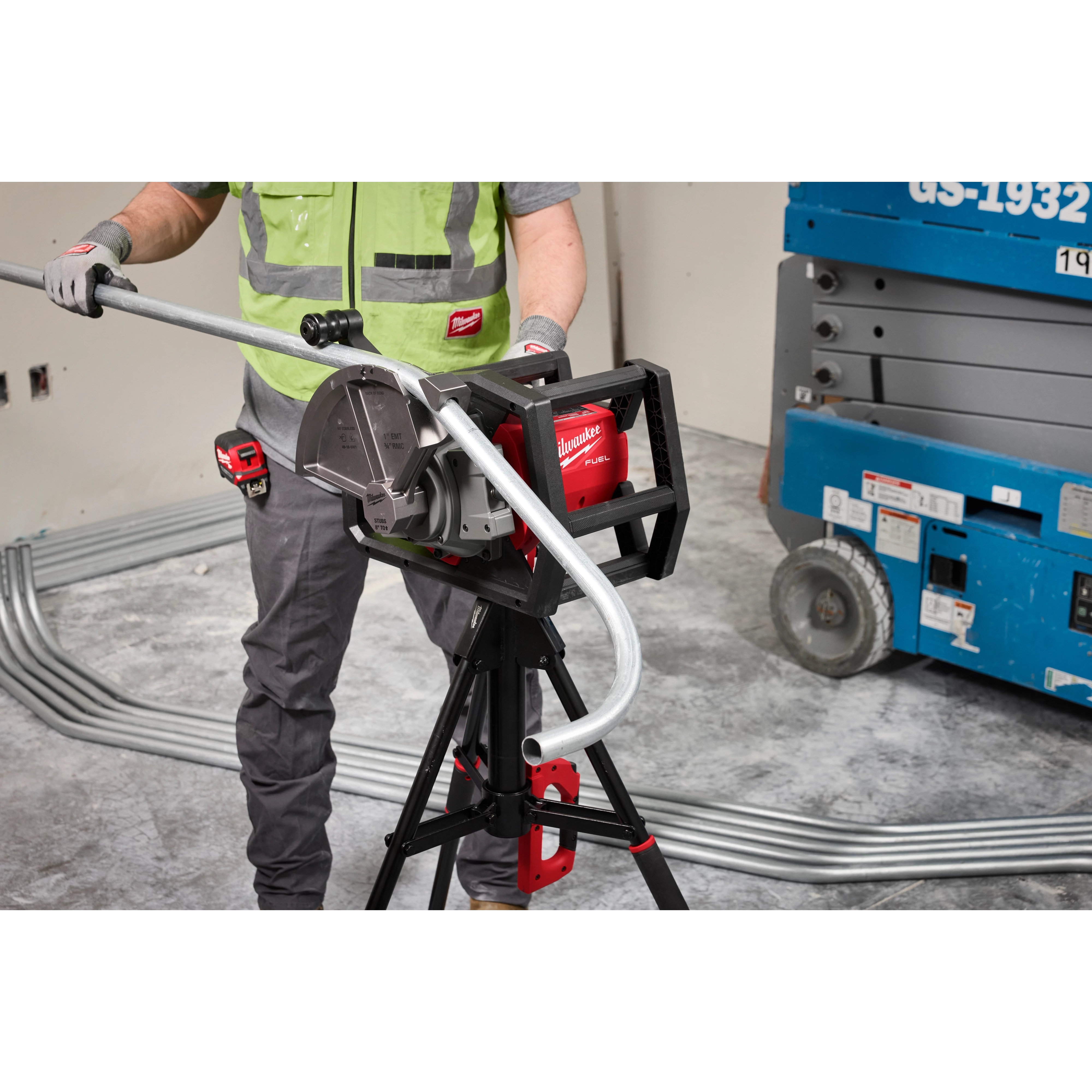 A worker operates the Milwaukee M18 FUEL Pipe Bender, which is mounted on a tripod stand. The device is bending metal piping in a construction setting. The worker wears a green safety vest and grey gloves, with additional pipes and construction equipment visible in the background.